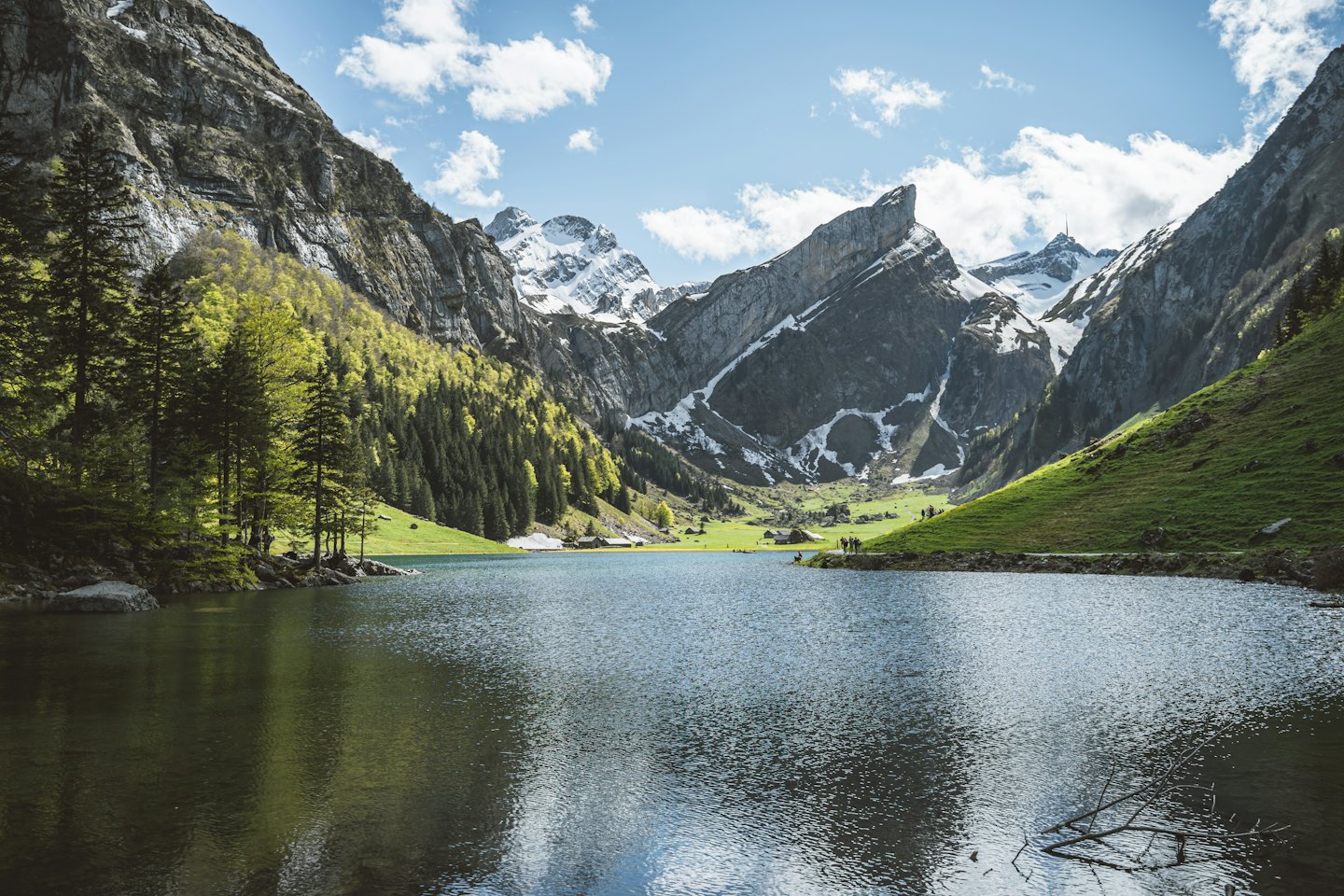 Persone passeggiano vicino a baite lungo un lago alpino panoramico.