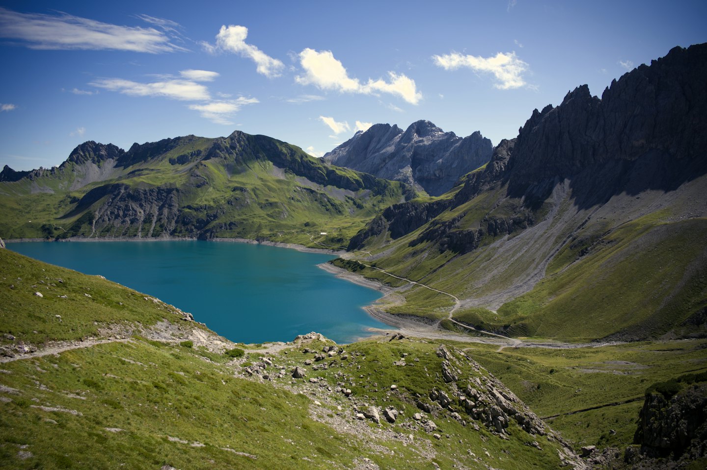 Lago alpino blu circondato da montagne con un sentiero sterrato.