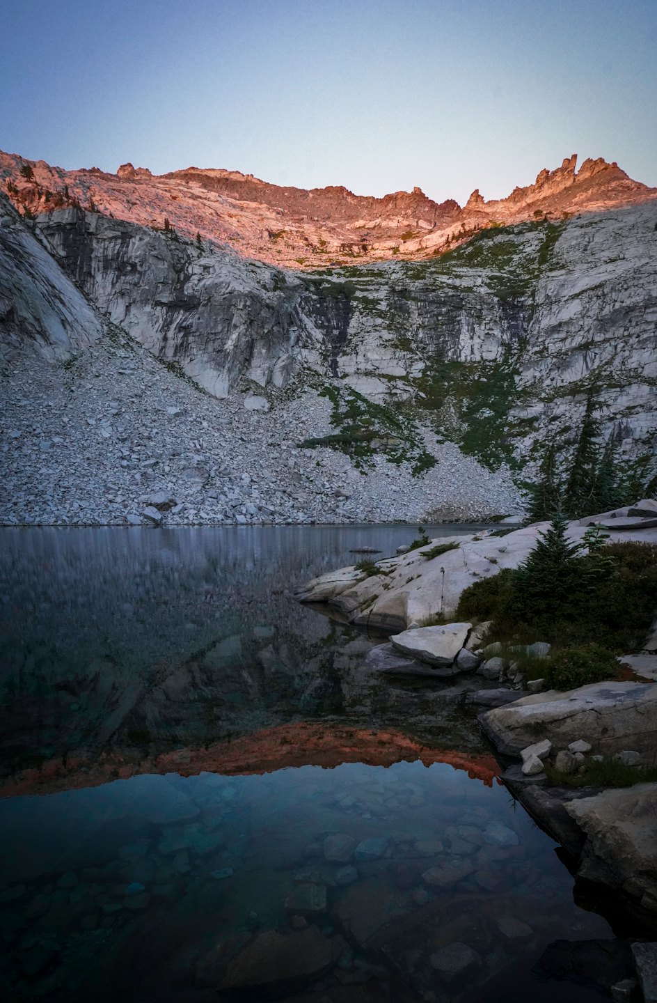 Lago alpino riflette montagne grigie sotto un cielo limpido.